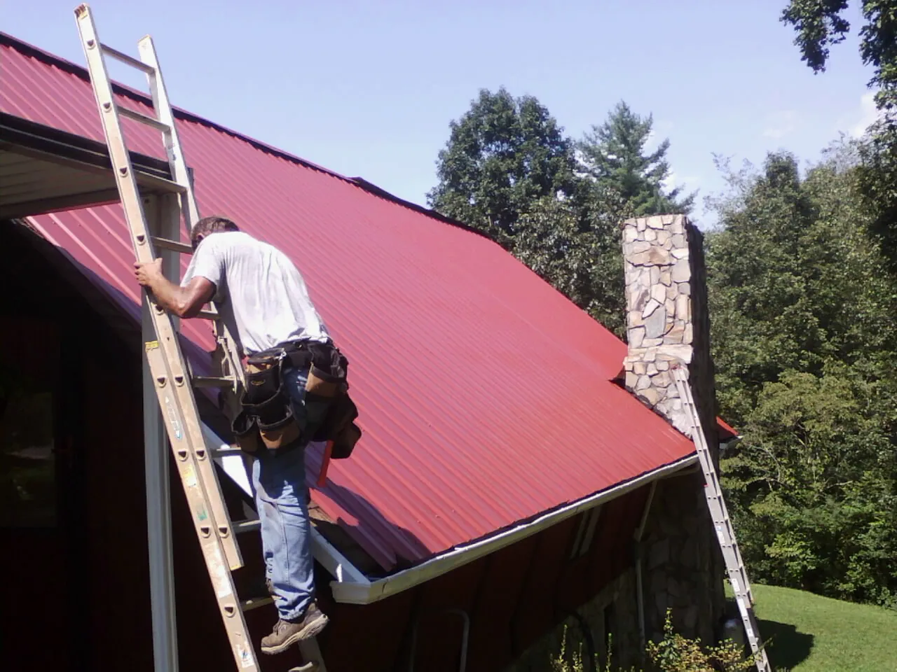 Roofer on a ladder beside a red metal roof and stone chimney — Soffit & Fascia Repair by Roofing Brothers in Calabasas